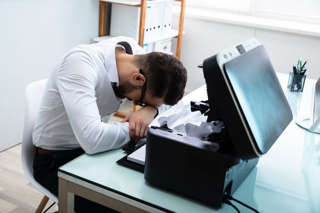 Frustrated office worker with their head down on a desk, sitting next to a malfunctioning printer or fax machine filled with crumpled paper