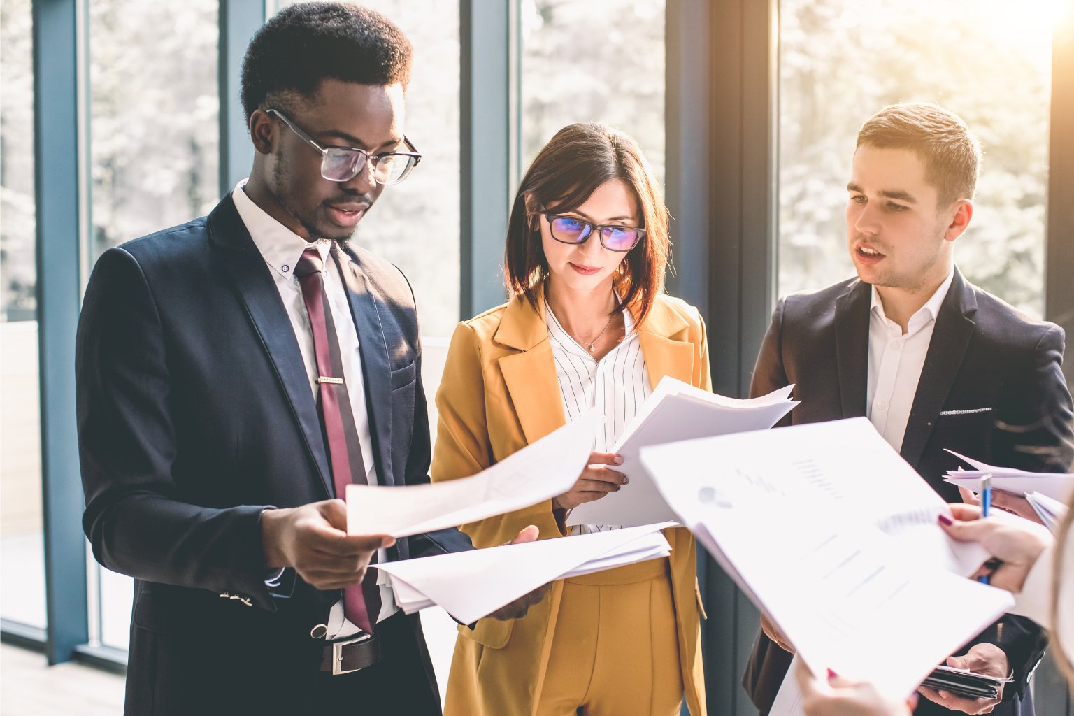 A group of business professionals review documents together in a bright, sunny office.
