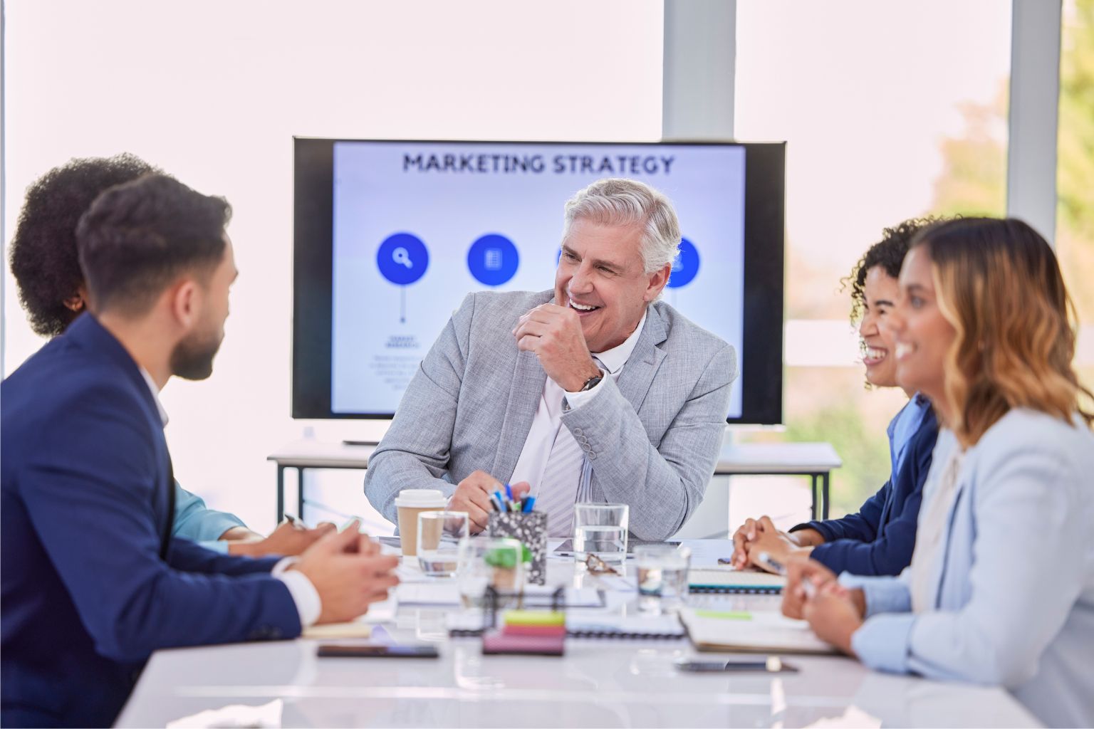 A group of five business professionals gathers together around a table to discuss marketing strategy. A screen in the background reads "Marketing Strategy" with blue icons.