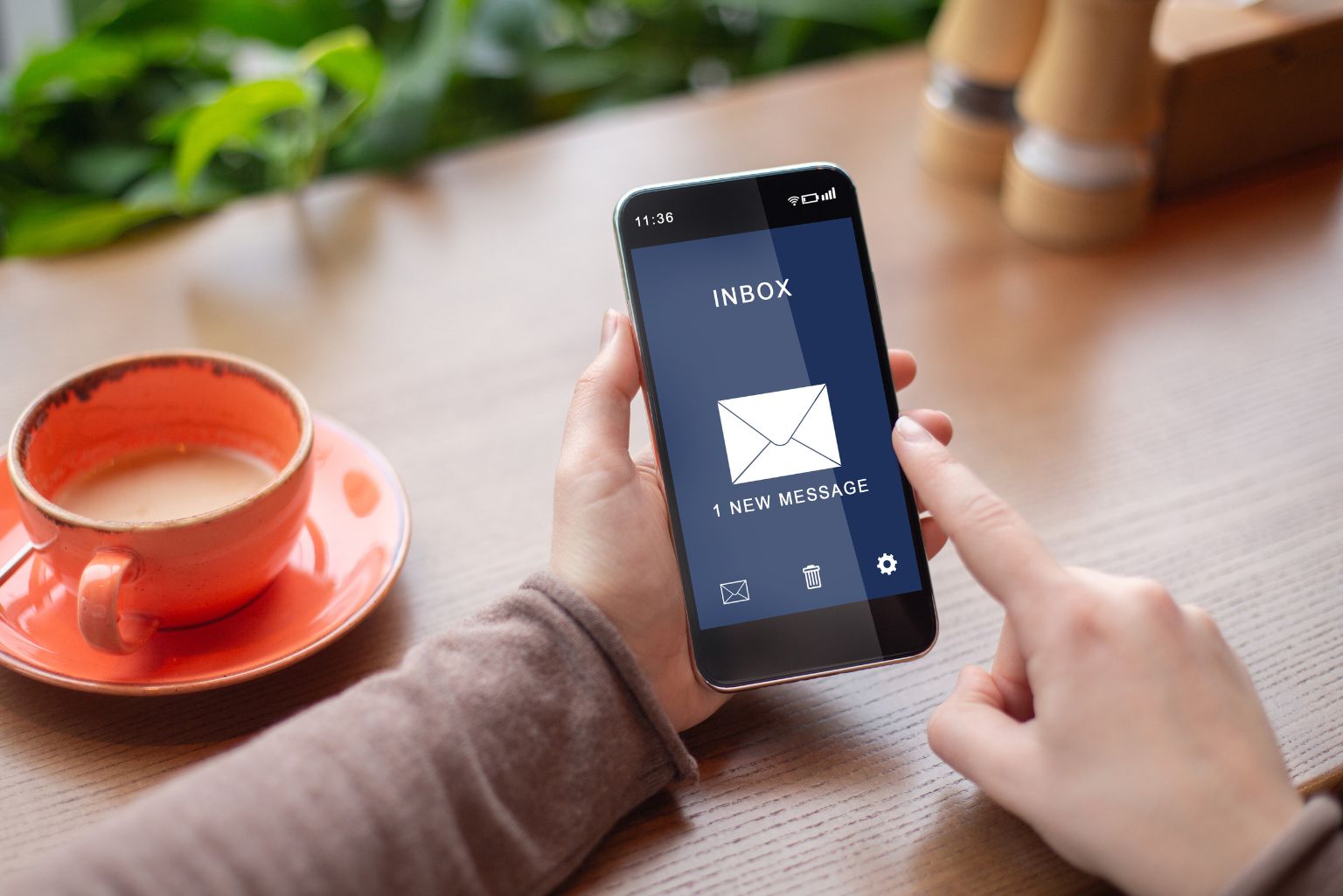 A pair of hands holding a smartphone over a desk with a cup of tea beside them. The screen of the smartphone shows the person checking their email inbox.