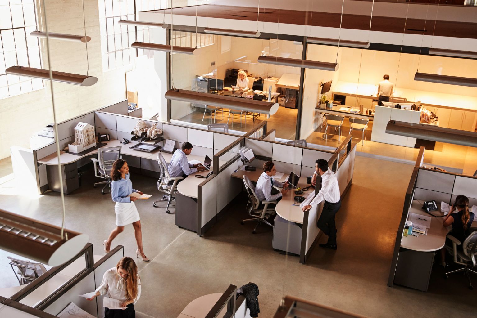 A high-angled view of a busy office building. Workers sit in cubicles on their computers while others walk through the office holding papers and folders.